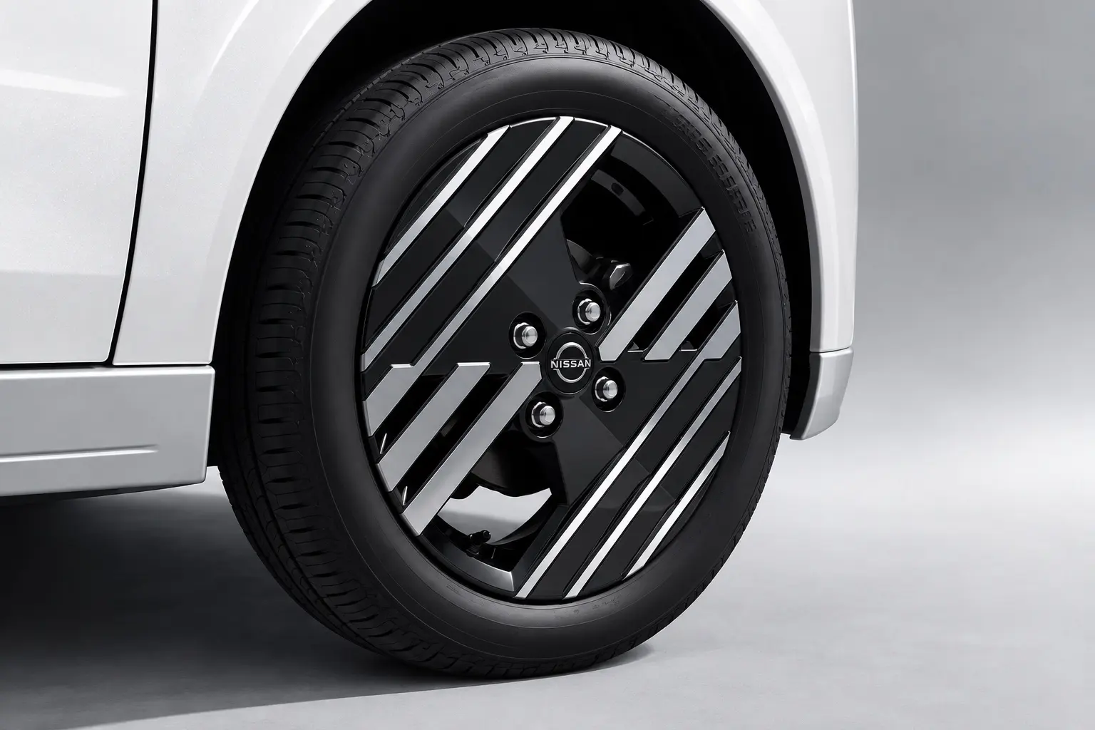 日産 サクラ(2026年モデルのG)の前輪まわりの寄りカット。グロスブラックの12本スポークで水引をイメージした15インチアルミホイール、165/55R15のタイヤ、ホワイトパールのボディが分かるカット。明るいスタジオ風の背景